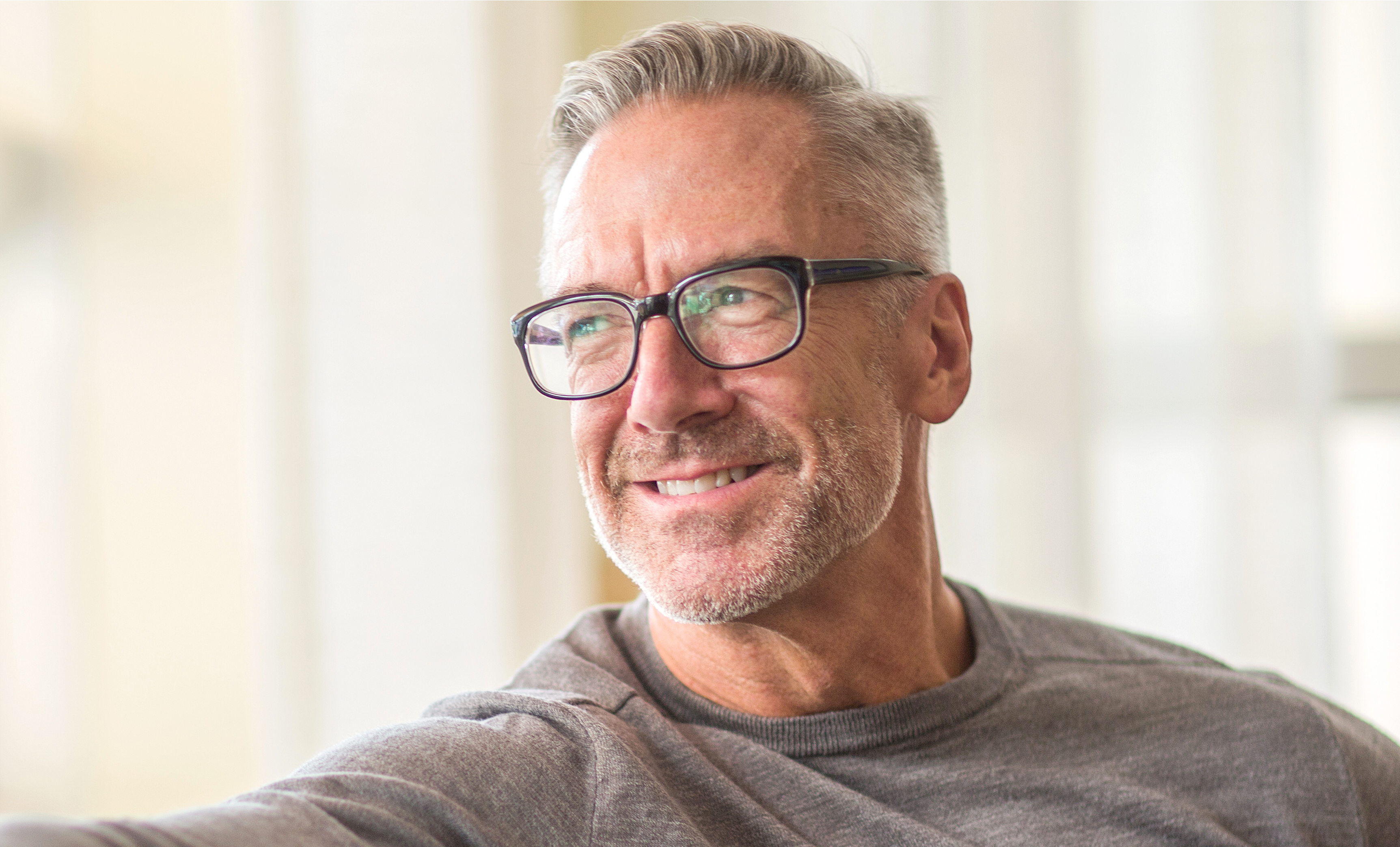 A male in his 50s wearing eye glasses and smiling while looking outside through a window.